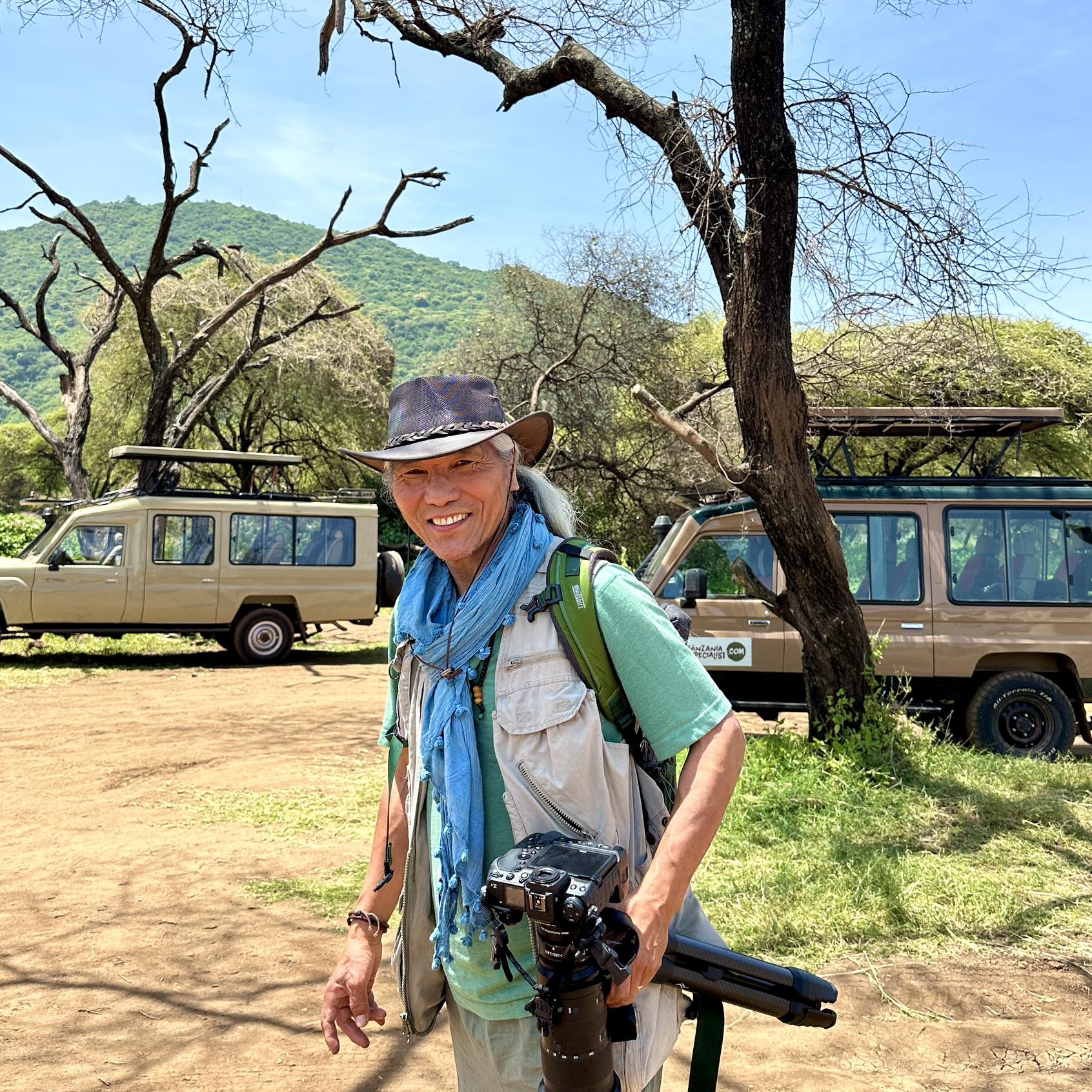 Sonam Zoksang on safari in Tanzania, wearing a hat and turquoise scarf, holding a camera and tripod, with acacia trees and safari vehicles behind him.